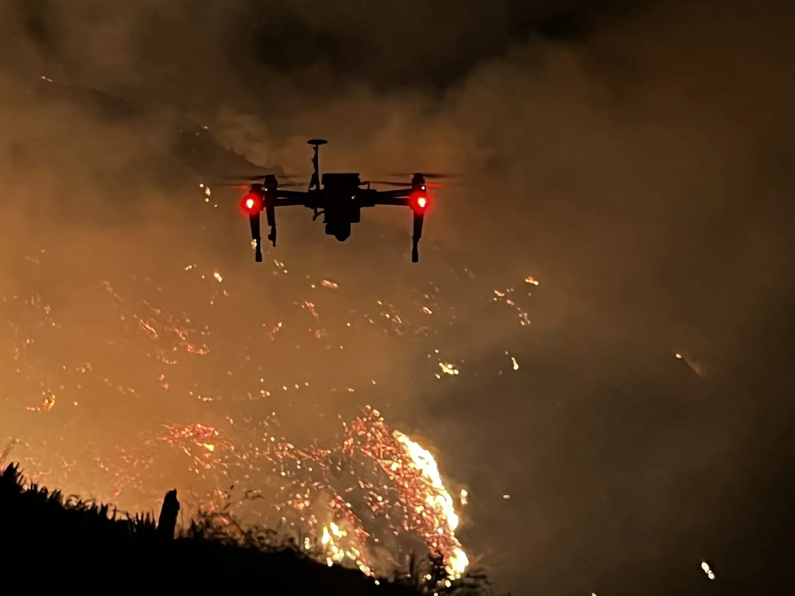 A drone hovering in smoke at night above a hillside of open flame and glowing embers, its red navigation lights lit.